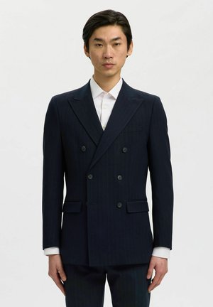 Hombre con traje cruzado de rayas finas azul marino oscuro y camisa blanca, de pie frente a un fondo blanco liso.