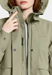 Vaaleanvihreä vedenpitävä takki, jossa on vetoketjullinen etuosa ja nepparit, säädettävä huppu ja pieni vetoketjullinen tasku rinnassa.