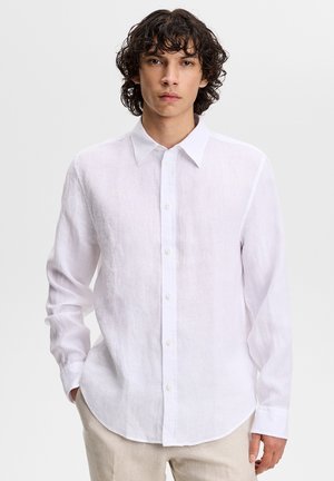 Giovane uomo con capelli ricci che indossa una camicia bianca a maniche lunghe con bottoni e pantaloni beige, in piedi davanti a uno sfondo neutro.