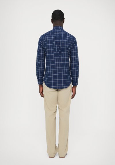 Temno modra karo srajca z dolgimi rokavi in ovratnikom na gumbe, v kombinaciji s svetlo bež hlačami. Srajca ima zaobljen spodnji rob in zadnjo gubo.