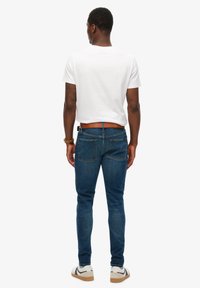 Męskie dopasowane jeansy z ciemnoniebieskiego denimu o gładkiej fakturze, z tylnymi kieszeniami i brązowym paskiem. Połączone z białym, krótkim T-shirtem i trampkami.