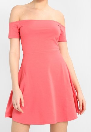 Femme portant une robe courte corail à épaules dénudées avec un corsage ajusté et une jupe évasée, sur un fond uni.