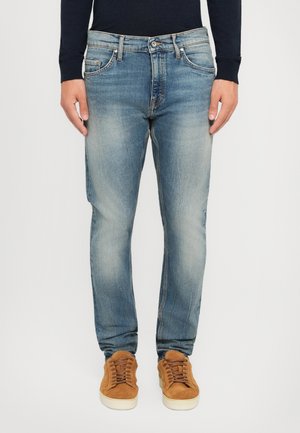 Man in vervaagde blauwe jeans, een zwart langemouwen shirt en bruin suède sneakers, staande tegen een eenvoudige witte achtergrond.