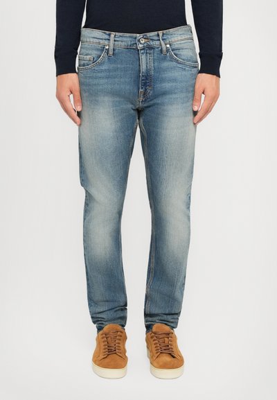 Hombre vistiendo jeans desgastados de color azul, una camisa negra de manga larga y zapatillas de ante marrones, de pie contra un fondo blanco liso.