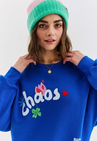 Modrá mikina s bielym, ružovým a zeleným grafickým motívom "chaos", obsahujúcim kvety a srdcové vzory. Doplnkovaná zelenou a ružovou pletenou čiapkou.