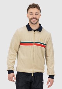 Beige Zip-Jacke mit einem marineblauen Kragen, ausgestattet mit einem rot-grünen Streifenband über der Brust und goldenen Hardware-Akzenten.