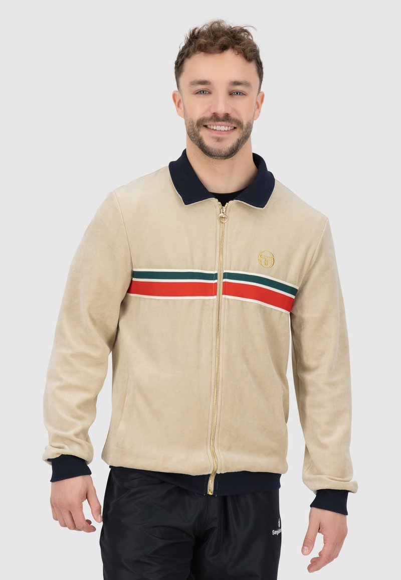 Beige Zip-Jacke mit einem marineblauen Kragen, ausgestattet mit einem rot-grünen Streifenband über der Brust und goldenen Hardware-Akzenten.