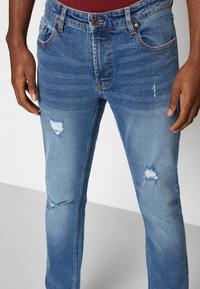 Blue denim jeans with a slim fit, featuring distressed areas, light fading, and copper-tone stitching. Five-pocket design with a button closure.