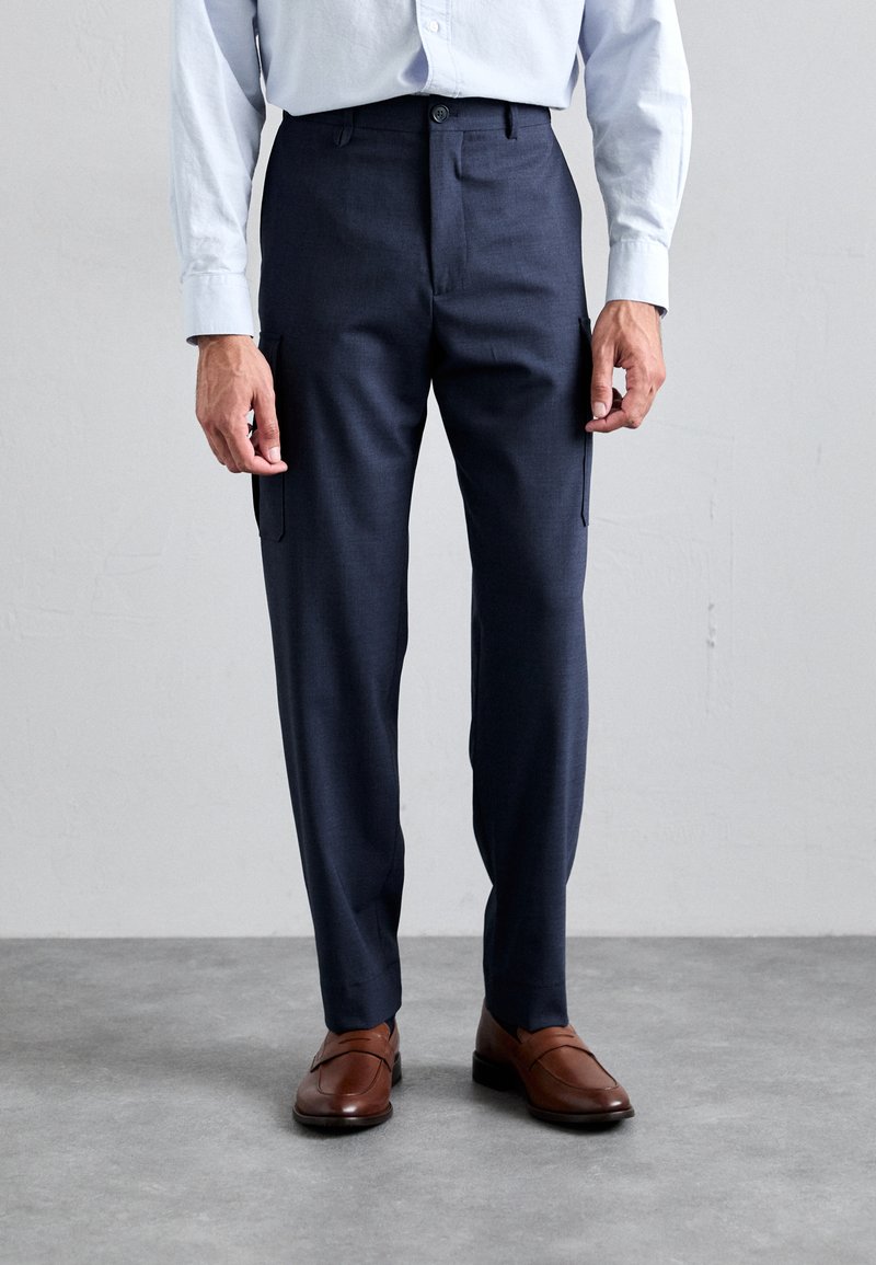 Pantalones de corte ajustado azul marino con bolsillos laterales, elaborados con una suave tela de mezcla de lana. Combinados con zapatos de cuero marrón.