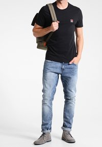 Černé tričko s krátkým rukávem a malým logem, světle modré džíny a šedé semišové boty. Modelka nese zelenou kabelku přes rameno.