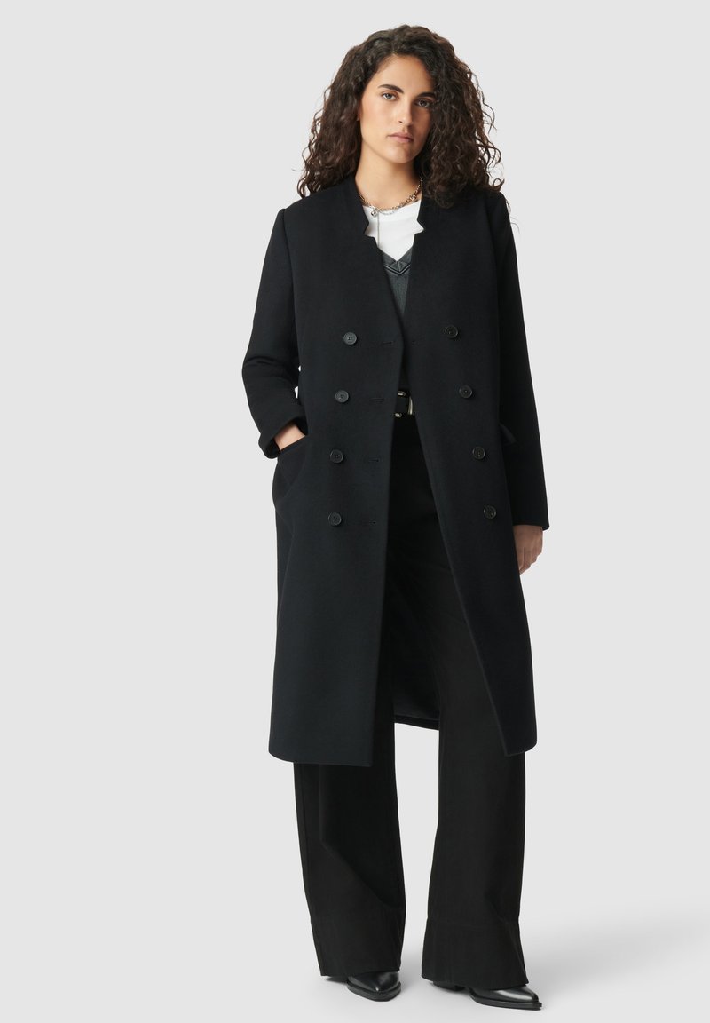 Vrouw met krullend, donker haar in een lange, zwarte, dubbele-breasted jas, zwarte, wijde broek en zwarte, puntige schoenen, staand tegen een grijze achtergrond.