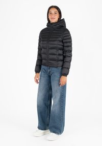 Zwarte pufferjack met een capuchon, voorzien van gestikte naden en een ritssluiting aan de voorkant. Gecombineerd met een wijde blauwe jeans en witte sneakers.
