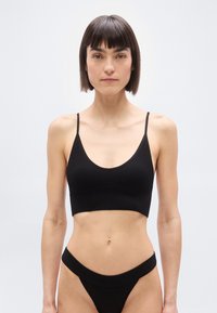 Λεπτή γυναίκα με κοντά σκούρα μαλλιά, φοράει μαύρο ριμπ crop top και ταιριαστό εσώρουχο με ψηλή μέση, στέκεται μπροστά σε απλό φωτεινό φόντο.