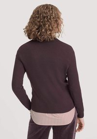 Dunkellila langärmeliger Pullover mit geripptem Saum, getragen über einem hellrosa und weißen gestreiften Shirt. Weicher Stoff und taillierte Passform.