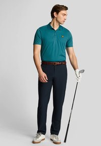 Teal polo shirt with a collar and embroidered logo, paired with dark navy pants. Accessories include a braided brown belt and white golf shoes.