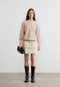 Γυναίκα που φοράει μπεζ φούτερ CK, ανοιχτό κίτρινη φούστα, μαύρες μπότες μέχρι το γόνατο, κρατώντας ένα μαύρο τσαντάκι, στέκεται μπροστά σε έναν απλό λευκό τοίχο.