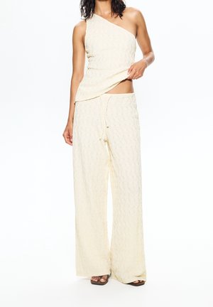 Vrouw draagt een crèmegekleurd, getextureerd een-schoudertopje en bijpassende wijduitlopende broek, houdt topje iets op om de buik bloot te geven.