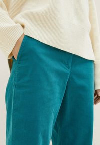 Pantalon en velours côtelé teal avec une texture lisse, doté de poches latérales, associé à un pull en maille côtelée beige clair. L'accent est mis sur le vêtement du bas.