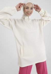 Pullover bianco oversize con colletto alto, half-zip e tasca laterale. Presenta maniche arricciate e una texture di tessuto morbido e liscio. Abbinato a pantaloni rosa.
