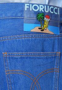 Plave traperice s narančastim šavovima, koje imaju oznaku marke Fiorucci s crtežom karikature sladoleda koji sjedi pored palme na otoku.