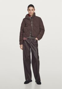 Braune gesteppte Jacke mit hohem Kragen, Reißverschluss, getragen über einem grauen Pullover, kombiniert mit weit geschnittenen, dunkelbraunen Lederhosen und schwarzem Schuhwerk.