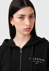 Melnas zipa kapuce ar mīkstu audumu. Tajā ir kapuce ar auklu un baltas burti uz krūtīm, kā arī metāla rāvējslēdzējs.