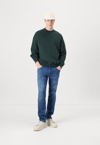Sudadera de cuello redondo verde oscuro, vaqueros azules y zapatillas crema. La sudadera tiene mangas anchas; los vaqueros son de corte ajustado. Gorro blanco en la cabeza.