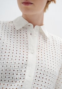 Blusa di cotone bianca con un motivo testurizzato in trama aperta. Presenta un colletto e chiusura con bottoni lungo la parte anteriore.