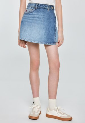 Osoba nosząca niebieską asymetryczną mini spódnicę z denimu, białe trampki i kremowe skarpetki na jednolitym tle.