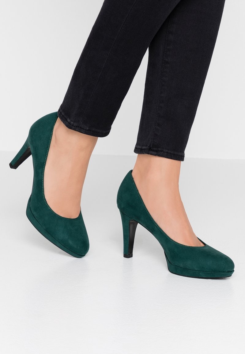 Personne portant des escarpins en daim vert foncé avec un pantalon noir sur un fond clair uni.