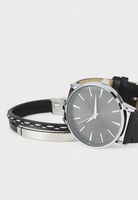 Montre ronde en argent avec bracelet en cuir noir, accents en argent et un cadran gris texturé avec de longs index des heures et une couronne centrale.
