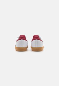 White sneakers with maroon accents, featuring a textured gum sole. The back has padded support and stitched detailing along the seam.