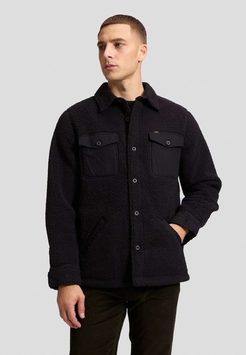 Homme portant une veste noire en laine texturée avec des poches poitrine boutonnées et des poches latérales sur un fond uni gris clair.