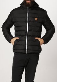 Chaqueta de plumas negra con capucha, con un cierre blanco, diseño acolchado horizontal y una pequeña etiqueta marrón en el área del pecho izquierdo.