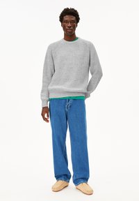 Grauer gerippter Pullover mit Rundhalsausschnitt, über einem grünen Shirt getragen. Kombiniert mit lockeren blauen Jeans und beigen Slippern. Einheitlicher weißer Hintergrund.
