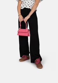 Mini borsa in finta pelle rosa con manico superiore, pantaloni neri a gamba larga e sneakers marroni con accenti e lacci rosa, su sfondo bianco.