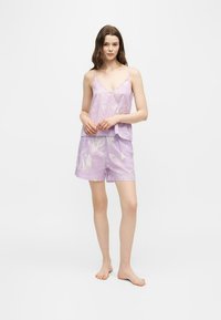 Femme debout pieds nus, portant un débardeur ample couleur lavande et un short assorti avec un léger motif de feuilles, sur fond blanc uni.