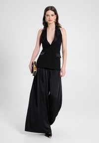 Schwarze taillierte Weste mit tiefem V-Ausschnitt, satinierten Akzenten, kombiniert mit weit geschnittenen schwarzen Hosen. Das Modell hält eine goldene Clutch und trägt spitze High Heels.