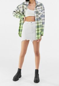 Modellen poserar i en grönrutigt överstor skjorta, vit crop-top och en nötig vit denimkjol, iförd svarta ankelboots.