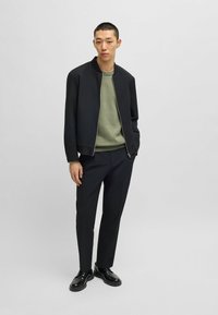 Schwarze Bomberjacke über einem hellgrünen Pullover, kombiniert mit schwarzen Hosen und schwarzen Schuhen, vor einem neutralen Hintergrund. Einfaches Design und Passform.