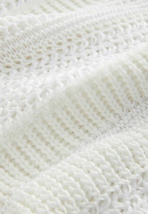 Balta adīta sega ar dažādām tekstūrām un rakstiem, izmantojot gan gludas, gan pūkainas daļas, radot slāņotu izskatu.