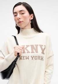 Maglione lavorato a maglia in crema con collare alto e logo "DKNY NEW YORK" in marrone. Il modello tiene una borsa in pelle nera, mettendo in mostra una texture liscia.