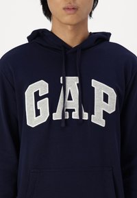 Sudadera con capucha de color azul marino fabricada en algodón, con grandes letras "GAP" en gris claro, cordón ajustable y un bolsillo frontal.
