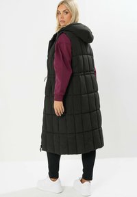 Svart vadderad väst med huva, med ett quiltat mönster, sidogömda dragkedjor och dragsko i midjan, som är tillsammans med en vinröd långärmad tröja.