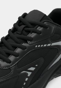 Μαύρο αθλητικό παπούτσι με υφή στο επάνω μέρος, κλείσιμο με κορδόνια, αντανακλαστικές λεπτομέρειες και μαλακή σόλα με κομψό σχέδιο.