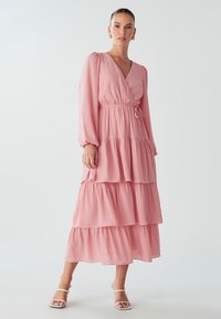 Růžové maxi šaty s vrstvením, dlouhými rukávy, hlubokým výstřihem do V a páskem v oblasti pasu. Vyrobeno z lehkého materiálu s plynulým střihem a hladkou texturou.