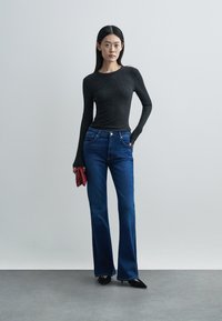 Top preto de manga comprida com malha canelada, combinado com jeans em denim azul, estilo flare. Modelo segura uma carteira vermelha; sapatos pretos de biqueira pontiaguda completam o visual.