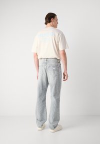 Világos bézs pamut póló kék márkajelzéssel a hátoldalon, világosszürke csíkos farmernadrággal párosítva. A modell fehér tornacipőt visel.