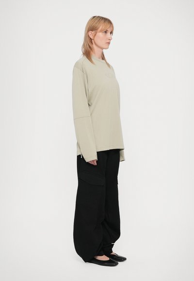 Cămașă lungă cu mânecă lungă de culoare verde deschis, cu textură, cu umeri lăsați, asociată cu pantaloni cargo negri, largi. Modelul stă în profil lateral, purtând încălțăminte plată.
