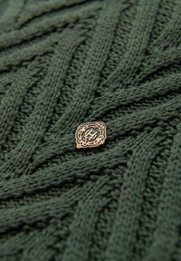 Sweter dziergany w kolorze leśnej zieleni, z teksturowanym wzorem. Ozdobiony złotą naszywką z misternym wzorem w okolicy środka.